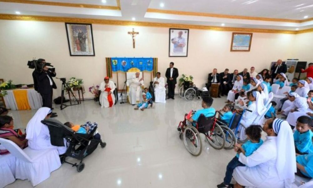 Fotografija prikazuje unutrašnjost prostorije gdje se odvija vjerski obred ili ceremonija. U središtu je par koji stoji ispred svećenika, a oko njih su prisutni gosti. Neki od gostiju su u invalidskim kolicima, a među prisutnima su i osobe u redovničkim odorama. Na zidu iza svećenika visi slika religijskog motiva. Prostorija je prostrana i dobro osvijetljena, a događaj snima kamera.