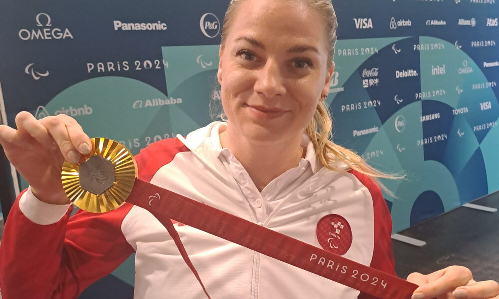 Fotografija prikazuje sportašicu u invalidskim kolicima koja drži zlatnu medalju osvojenom na Paraolimpijskim igrama u Parizu 2024. godine. Na sportašici je službena odjeća hrvatske reprezentacije, a u pozadini se vidi plakat s logotipima sponzora i natpisom "Paris 2024".