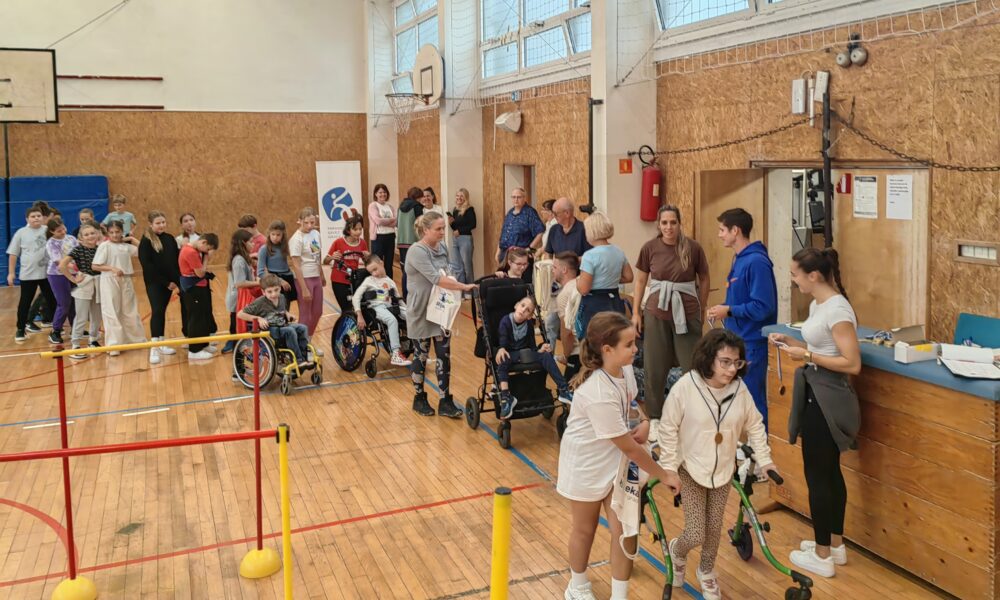 Na fotografiji su prikazana djeca u sportskoj dvorani. Na parketu sportske dvorane nalaze se rekviziti za vježbanje. Fotografija prikazuje dodjelu medalja djeci koja su sudjelovala na natjecanju. U daljini se nazire koš. plava strunjača i baner od organizatora natjecanja.