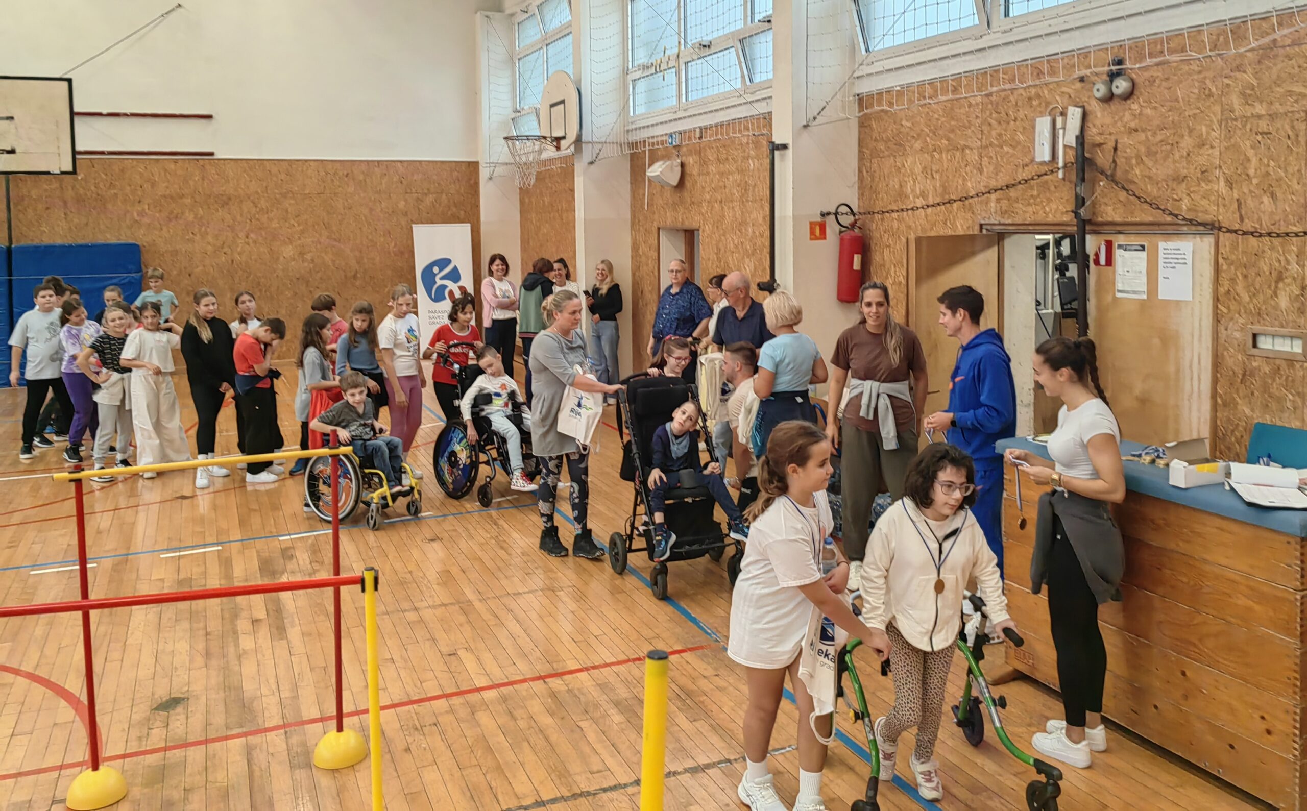 Na fotografiji su prikazana djeca u sportskoj dvorani. Na parketu sportske dvorane nalaze se rekviziti za vježbanje. Fotografija prikazuje dodjelu medalja djeci koja su sudjelovala na natjecanju. U daljini se nazire koš. plava strunjača i baner od organizatora natjecanja.