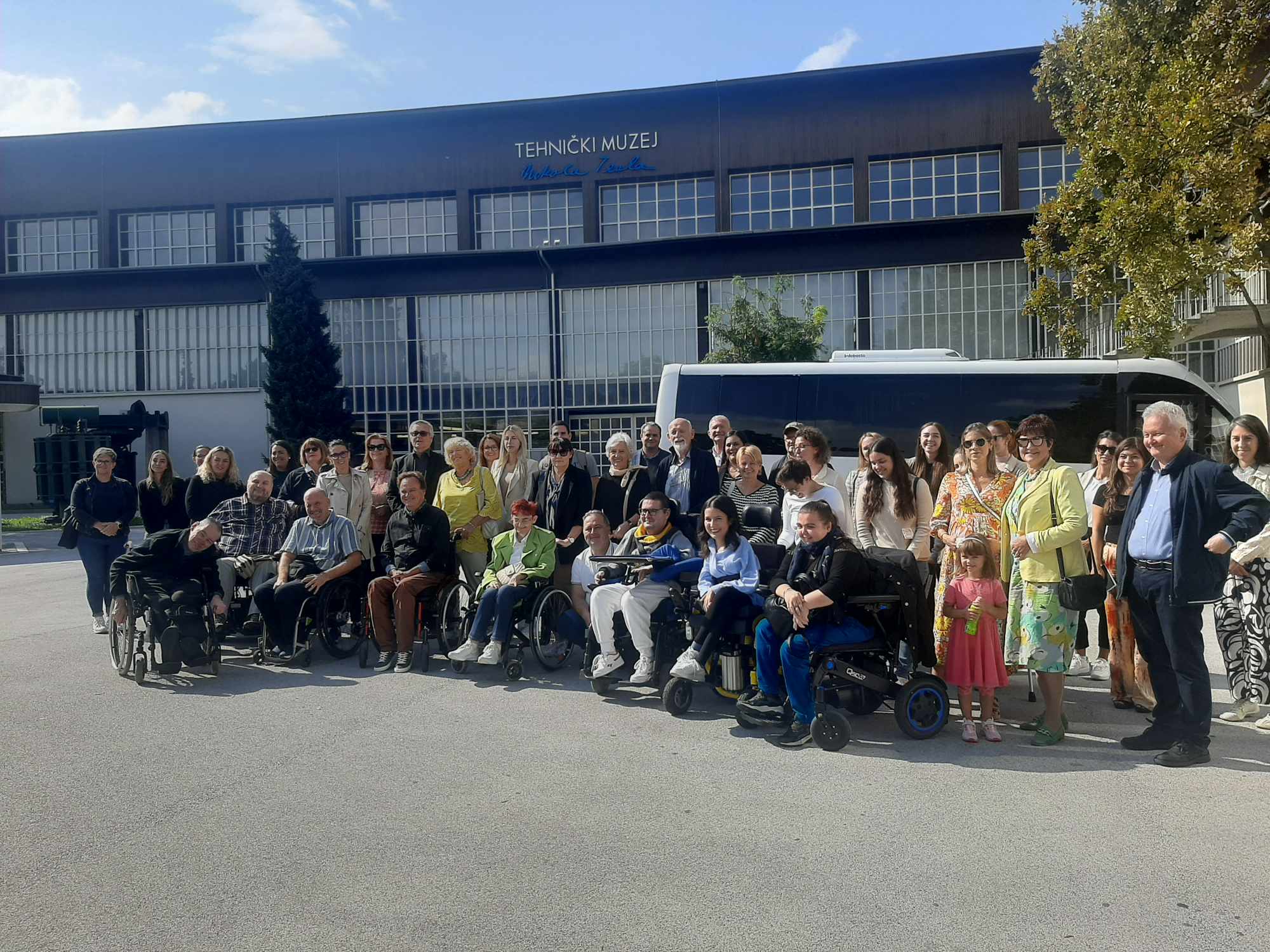 Fotografija prikazuje više ljudi ispred Tehničkog muzeja Nikole Tesle u Zagrebu, kako to sugerira plavi natpis na vrhu zgrade. Neki ljudi stoje, a neki su u invalidskim kolicima. Iza ljudi vidi se parkiran bijeli autobus.