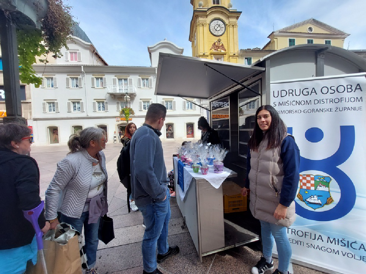 Skupina ljudi stoji pored štanda na otvorenom. Na štandu piše "Udruga osoba s mišićnom distrofijom Primorsko-goranske županije". Jedna žena se nasmiješeno obraća kameri, dok ostali ljudi gledaju izložene proizvode na štandu. Iza njih je trg i zgrada sa žutim tornjem u pozadini.