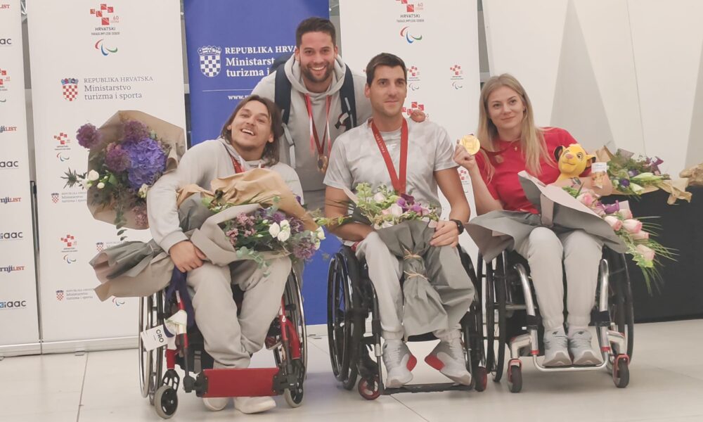 Slika prikazuje četvero hrvatskih paraolimpijaca (troje u invalidskim kolicima) s medaljama oko vrata. Svi drže velike bukete cvijeća i smiješe se kameri. Pozadina uključuje plakat s logom Ministarstva turizma i sporta Republike Hrvatske.