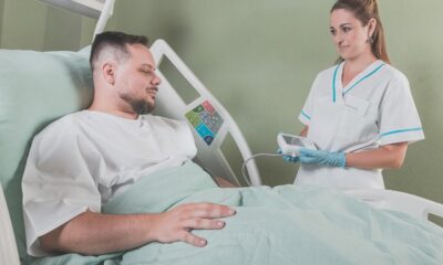 Na fotografiji je prikazana scena u bolničkoj sobi. Osoba u bijeloj medicinskoj uniformi stoji pored kreveta i drži neki uređaj, dok druga osoba leži na krevetu s podignutom glavom i izgleda kao da je pacijent. Oboje imaju zamagljena lica. Scena može biti zanimljiva ili relevantna jer prikazuje interakciju između medicinskog osoblja i pacijenta, što može ukazivati na pružanje zdravstvene skrbi ili medicinski postupak.