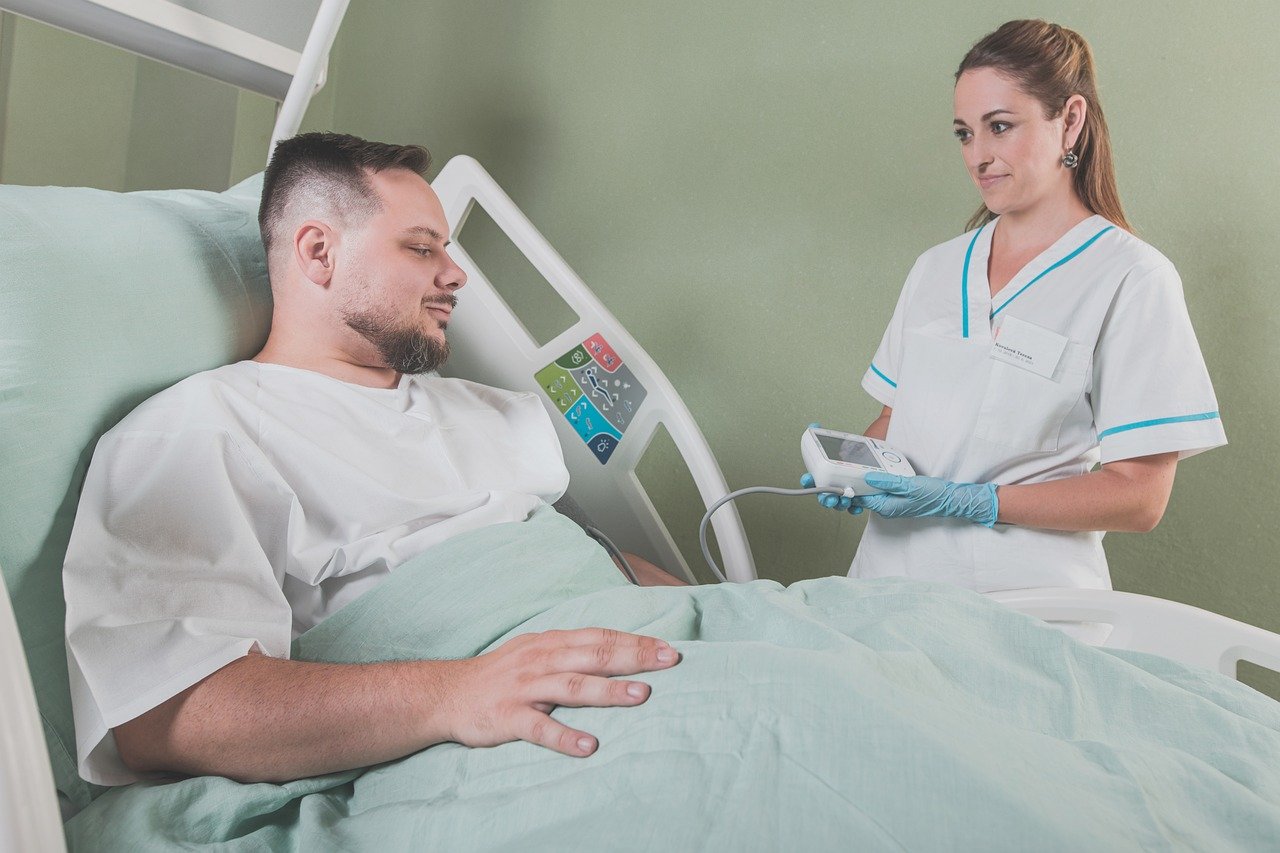 Na fotografiji je prikazana scena u bolničkoj sobi. Osoba u bijeloj medicinskoj uniformi stoji pored kreveta i drži neki uređaj, dok druga osoba leži na krevetu s podignutom glavom i izgleda kao da je pacijent. Oboje imaju zamagljena lica. Scena može biti zanimljiva ili relevantna jer prikazuje interakciju između medicinskog osoblja i pacijenta, što može ukazivati na pružanje zdravstvene skrbi ili medicinski postupak.