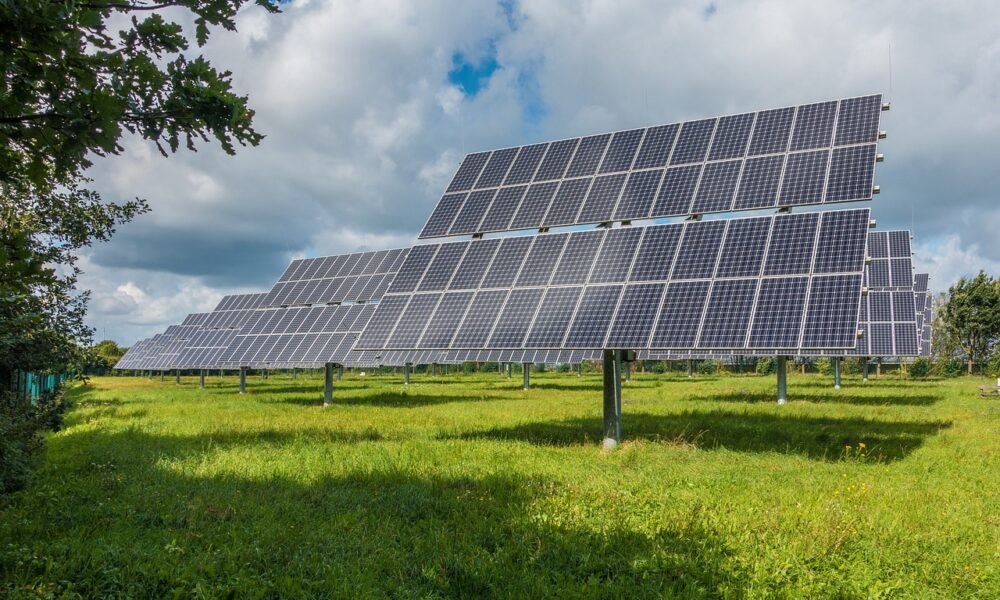 Veliki sustav solarnih panela postavljen na zelenom polju pod vedrim nebom s oblacima. Paneli su organizirani u redove i okrenuti prema suncu, što ukazuje na korištenje solarne energije.