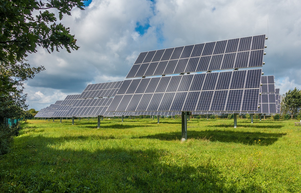 Veliki sustav solarnih panela postavljen na zelenom polju pod vedrim nebom s oblacima. Paneli su organizirani u redove i okrenuti prema suncu, što ukazuje na korištenje solarne energije.