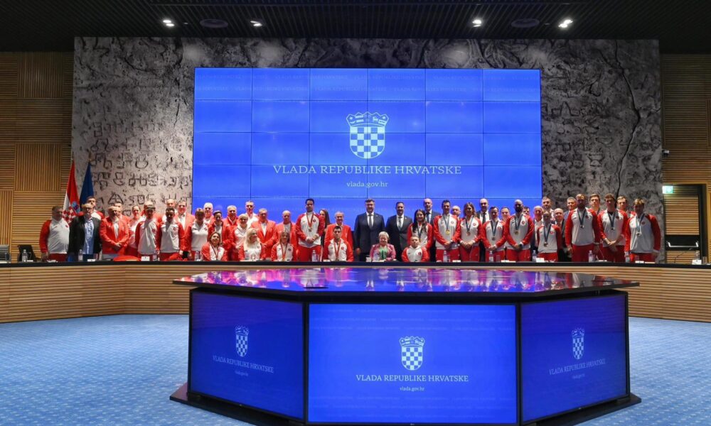 Na fotografiji je prikazano više ljudi u crveno bijelim trenirkama, a neki su i u sakoima. Većina ljudi stoji, a neki su i u kolicima. Neki imaju medalje oko vrata. Na zidu je plavi ekran na kojem piše 'Vlada Republike Hrvatske' i 'Vlada: gov.hr' Vjerojatno se radi o prijamu kod predsjednika ili premjera.