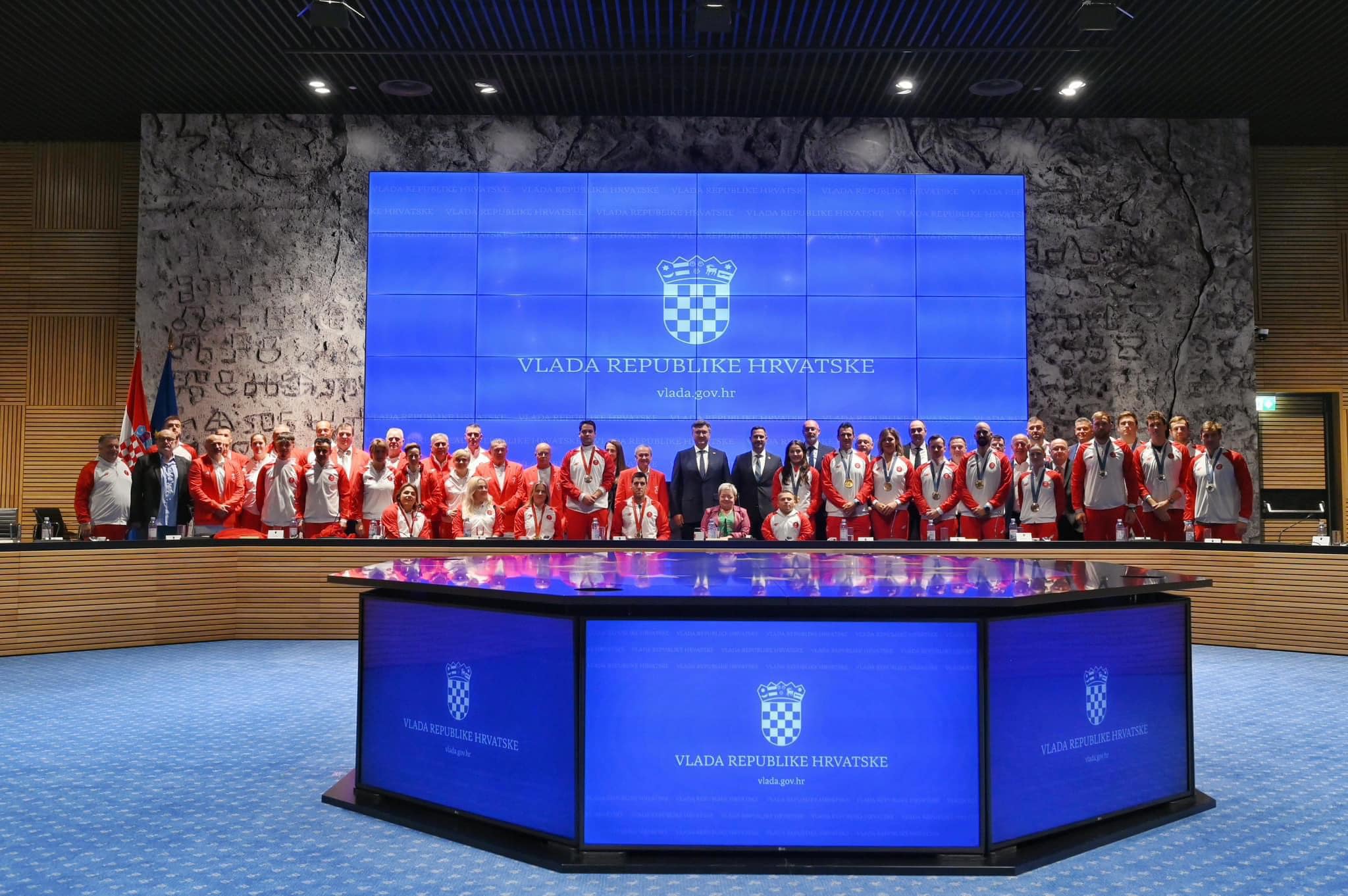 Na fotografiji je prikazano više ljudi u crveno bijelim trenirkama, a neki su i u sakoima. Većina ljudi stoji, a neki su i u kolicima. Neki imaju medalje oko vrata. Na zidu je plavi ekran na kojem piše 'Vlada Republike Hrvatske' i 'Vlada: gov.hr' Vjerojatno se radi o prijamu kod predsjednika ili premjera.