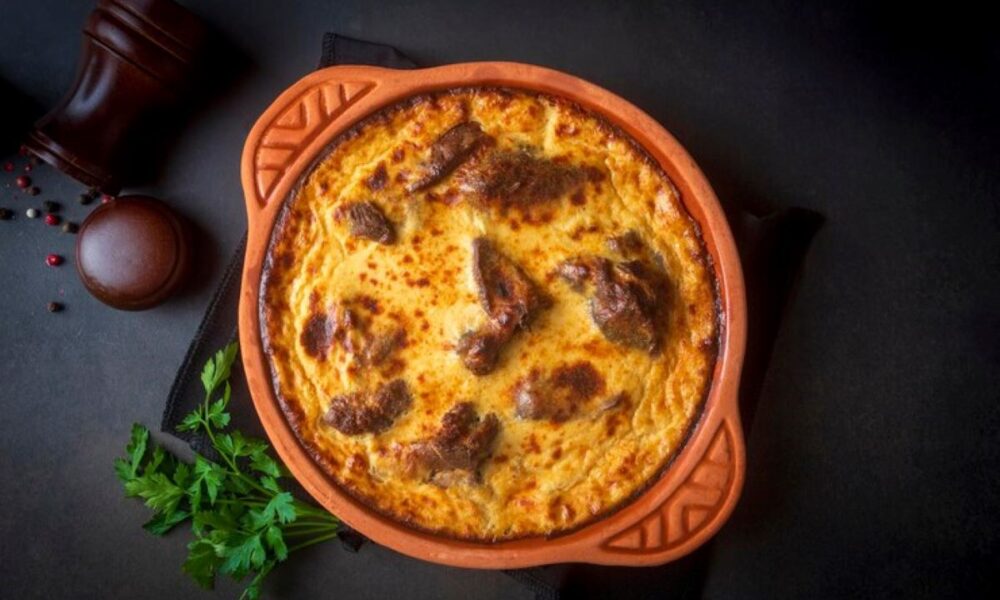 Na fotografiji je prikazano pečeno jelo u terakota posudi na tamnoj podlozi. Jelo izgleda kao zapečena tjestenina ili sličan gratinirani obrok, sa zlatno-smeđom korom na površini. Uz posudu se nalazi grančica peršina i mlinac za papar, što ukazuje na to da je jelo vjerojatno začinjeno i spremno za serviranje. Ova fotografija bi mogla biti zanimljiva ili relevantna zbog prikaza gotovog kulinarskog jela koje može privući pažnju ljubitelja hrane ili onih koji traže inspiraciju za kuhanje.