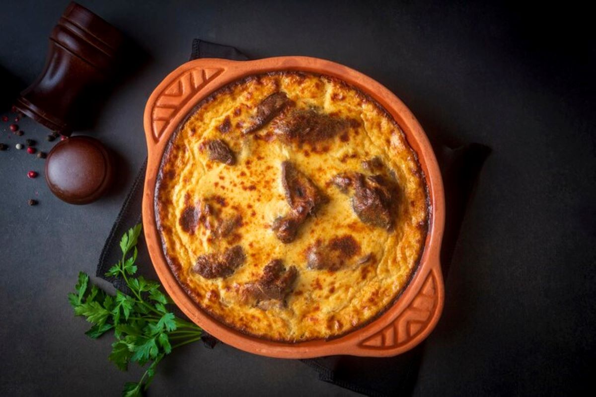 Na fotografiji je prikazano pečeno jelo u terakota posudi na tamnoj podlozi. Jelo izgleda kao zapečena tjestenina ili sličan gratinirani obrok, sa zlatno-smeđom korom na površini. Uz posudu se nalazi grančica peršina i mlinac za papar, što ukazuje na to da je jelo vjerojatno začinjeno i spremno za serviranje. Ova fotografija bi mogla biti zanimljiva ili relevantna zbog prikaza gotovog kulinarskog jela koje može privući pažnju ljubitelja hrane ili onih koji traže inspiraciju za kuhanje.