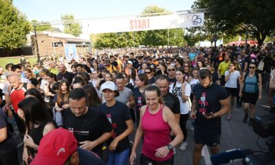 Velika skupina trkača okupljena na startu utrke na otvorenom. Ljudi su u sportskoj odjeći i pripremaju se za početak utrke. Iznad njih je natpis "START", a okolina je zelena s drvećem u pozadini. Atmosfera je energična i dinamična.