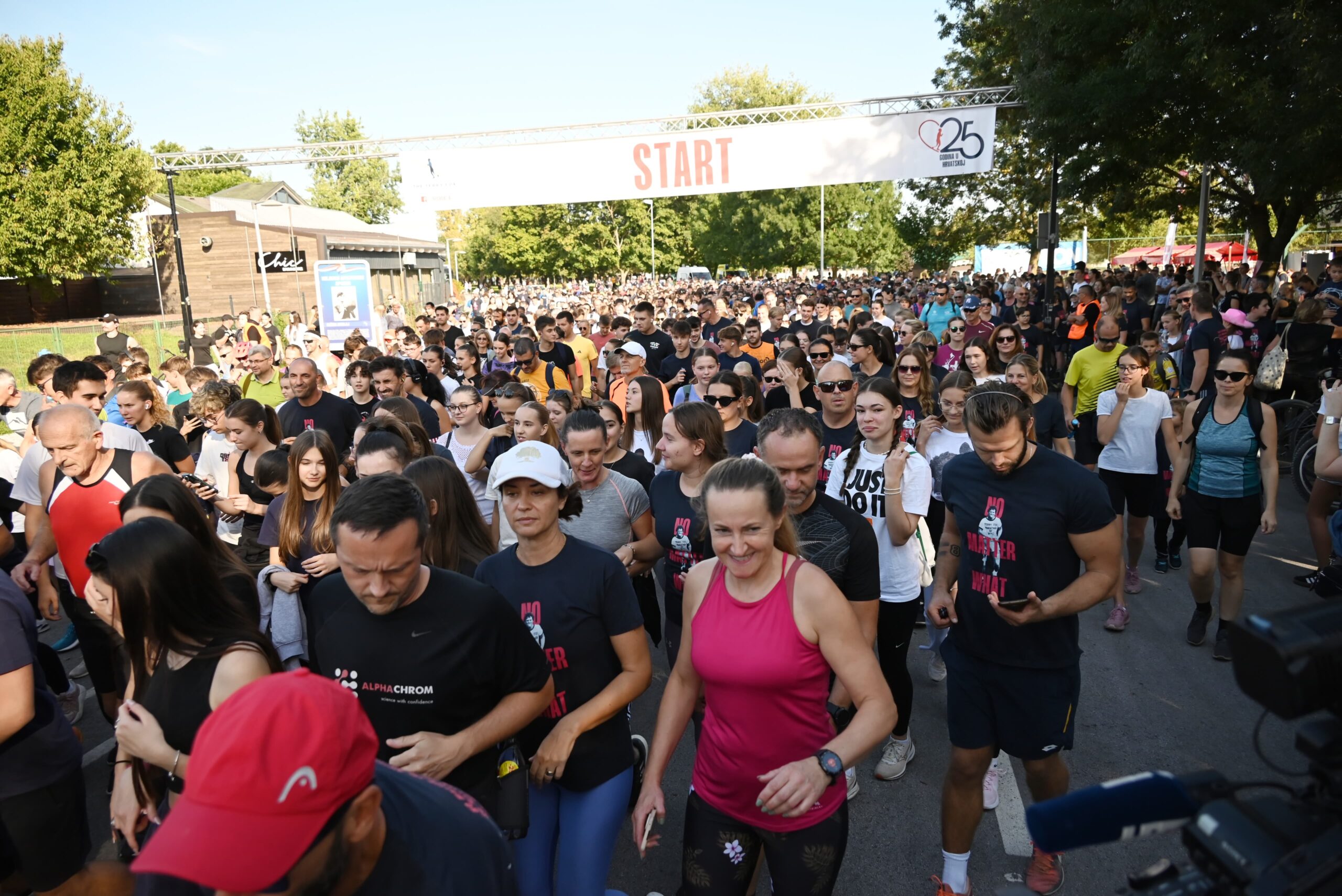 Velika skupina trkača okupljena na startu utrke na otvorenom. Ljudi su u sportskoj odjeći i pripremaju se za početak utrke. Iznad njih je natpis "START", a okolina je zelena s drvećem u pozadini. Atmosfera je energična i dinamična.