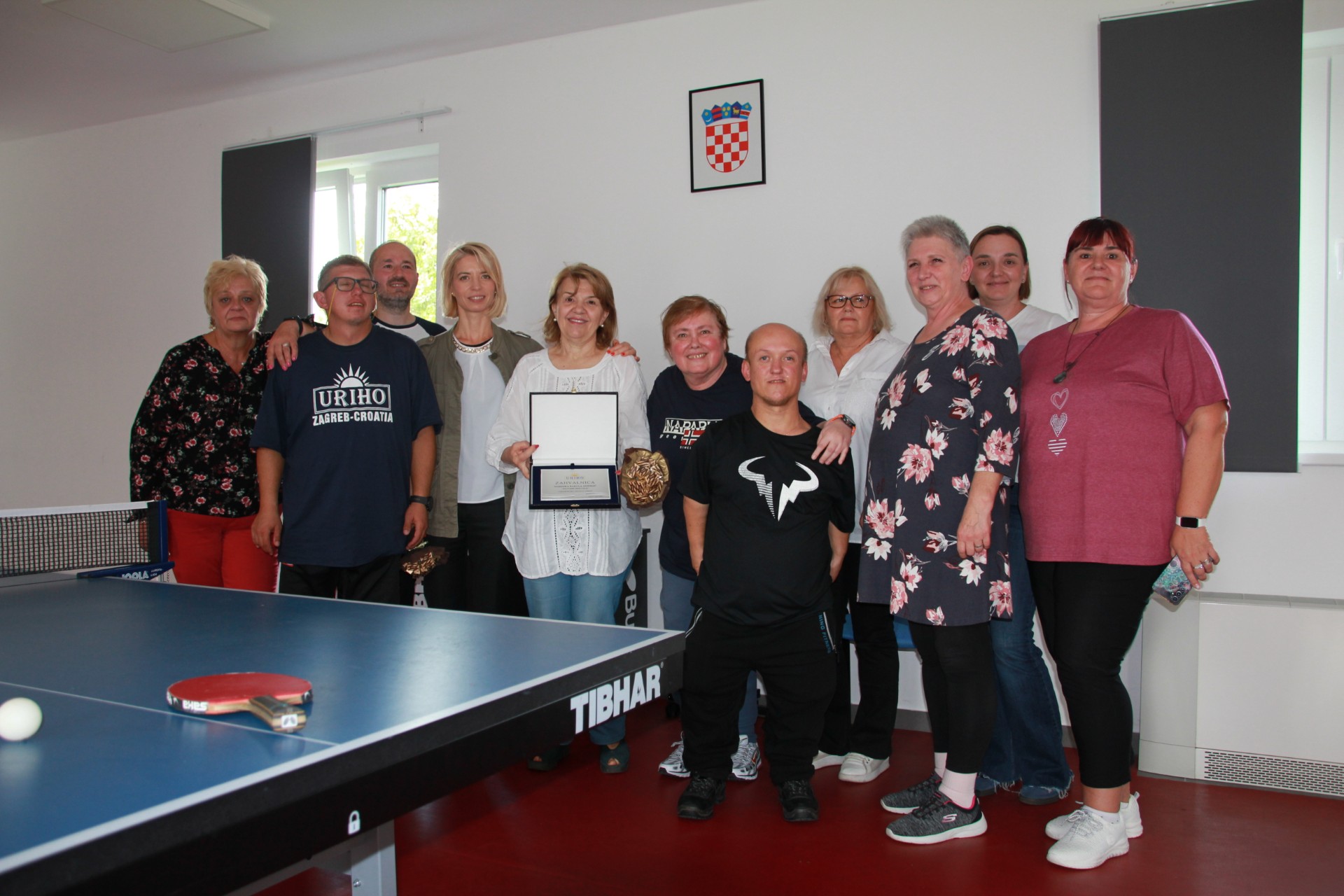 Na fotografiji je nekoliko osoba. Žena u sredini drži zahvalnicu. Fotografija je nastala u zatvorenom prostoru, a na njoj se vidi stol za stolni tenis na kojem se nalaze crveni stolnoteniski reket i bijela loptica. Iza ljudi nalazi se i slika s hrvatskim grbom.