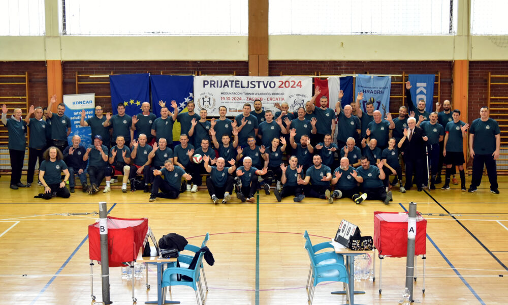 Grupna fotografija snimljena u sportskoj dvorani, na kojoj su deseci sudionika, većinom muškarci, svi odjeveni u tamnozelene majice, u dvorani za sjedeću odbojku. Svi sudionici poziraju s osmijehom i podignutim rukama, pokazujući znak prijateljstva. Iza njih je veliki transparent s natpisom "Prijateljstvo 2024" i informacijama o međunarodnom turniru u sjedećoj odbojci, koji se održava u Zagrebu, 19.10.2024. Na transparentu su logotipi organizatora, a sa strane su zastave Europske unije i drugih partnera. U prednjem planu vidi se odbojkaška mreža, stol za suce, stolice i oprema za sjedeću odbojku.