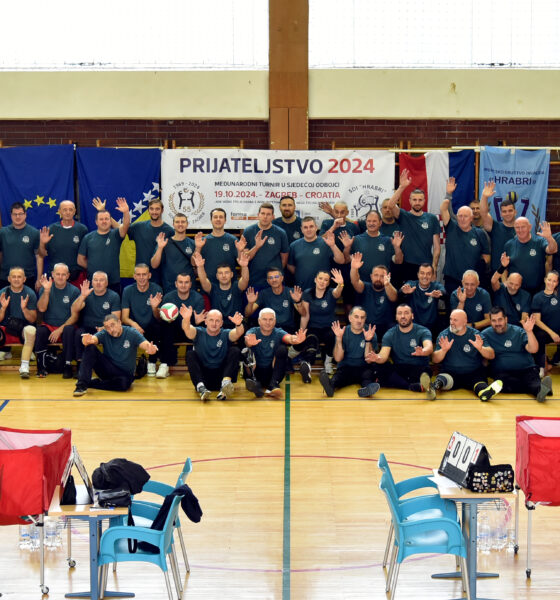 Grupna fotografija snimljena u sportskoj dvorani, na kojoj su deseci sudionika, većinom muškarci, svi odjeveni u tamnozelene majice, u dvorani za sjedeću odbojku. Svi sudionici poziraju s osmijehom i podignutim rukama, pokazujući znak prijateljstva. Iza njih je veliki transparent s natpisom "Prijateljstvo 2024" i informacijama o međunarodnom turniru u sjedećoj odbojci, koji se održava u Zagrebu, 19.10.2024. Na transparentu su logotipi organizatora, a sa strane su zastave Europske unije i drugih partnera. U prednjem planu vidi se odbojkaška mreža, stol za suce, stolice i oprema za sjedeću odbojku.