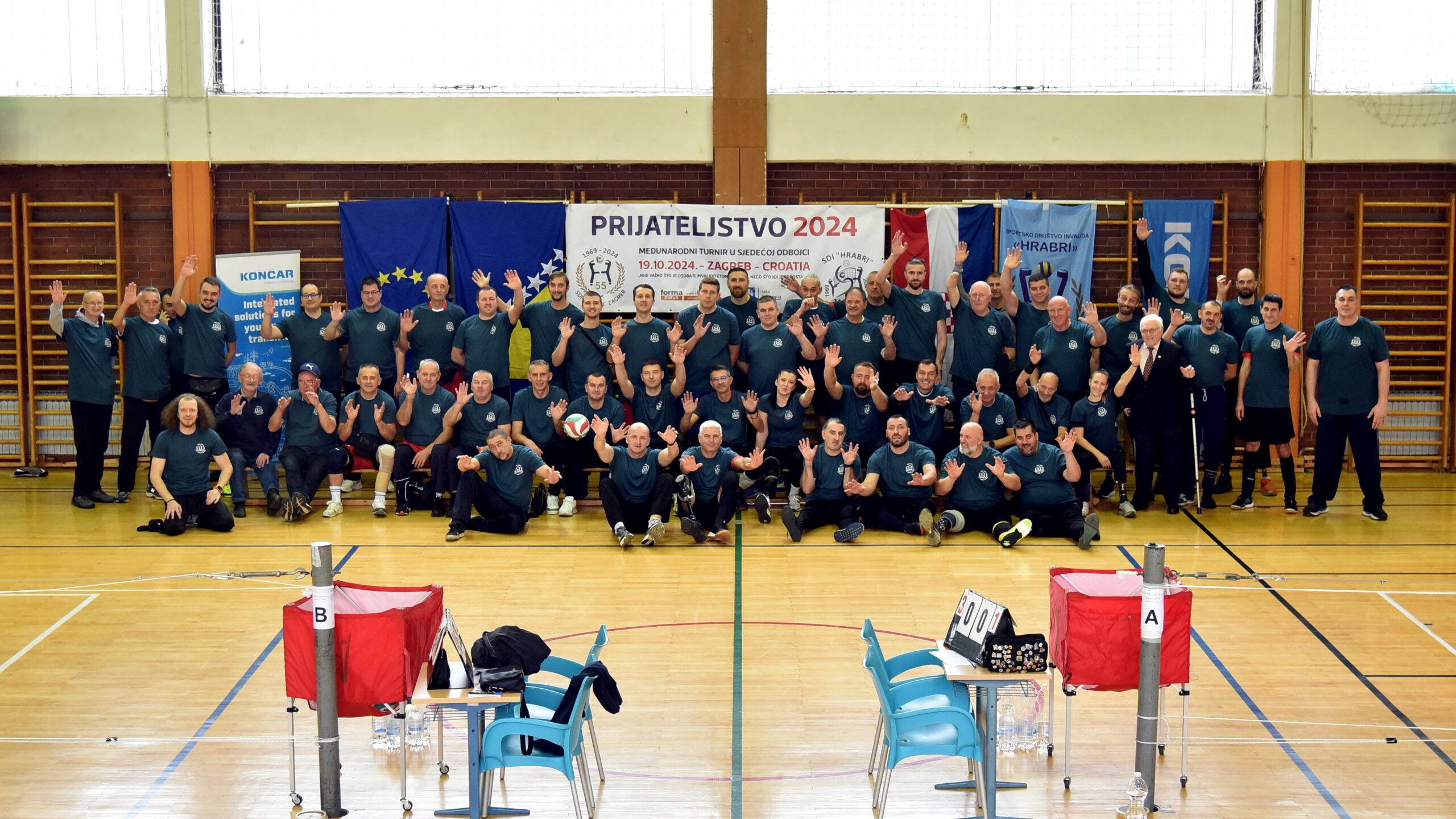 Grupna fotografija snimljena u sportskoj dvorani, na kojoj su deseci sudionika, većinom muškarci, svi odjeveni u tamnozelene majice, u dvorani za sjedeću odbojku. Svi sudionici poziraju s osmijehom i podignutim rukama, pokazujući znak prijateljstva. Iza njih je veliki transparent s natpisom "Prijateljstvo 2024" i informacijama o međunarodnom turniru u sjedećoj odbojci, koji se održava u Zagrebu, 19.10.2024. Na transparentu su logotipi organizatora, a sa strane su zastave Europske unije i drugih partnera. U prednjem planu vidi se odbojkaška mreža, stol za suce, stolice i oprema za sjedeću odbojku.