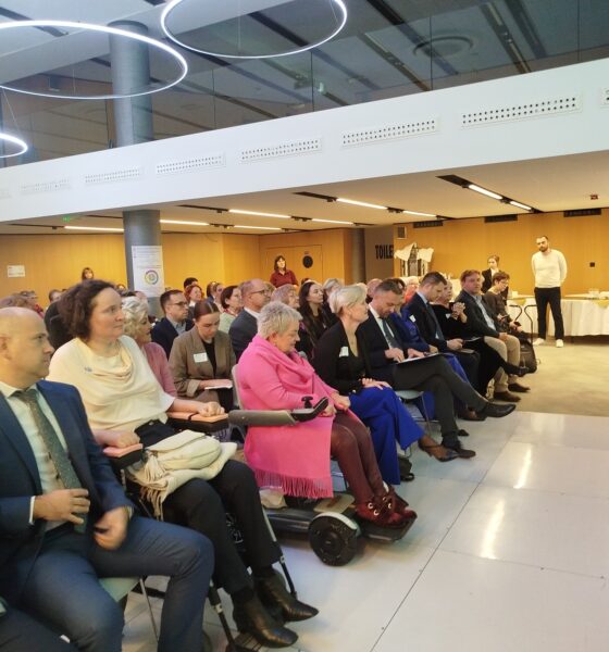 Fotografija prikazuje skupinu ljudi koji sjede u konferencijskoj dvorani. Svi su usmjereni prema govorniku koji stoji na pozornici s mikrofonom. U pozadini se nalazi pult s natpisima, a prostor je osvijetljen umjetnim svjetlom. Čini se da se radi o nekom događaju poput konferencije ili predavanja.