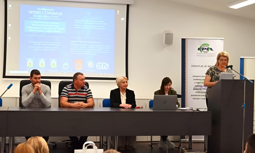 Panel od četvero ljudi koji sjede za stolom ispred ekrana s prezentacijom. Tema konferencije je "Sport i zdravlje", a jedan od govornika, žena, drži govor s govornice, dok ostali panelisti sjede i slušaju.