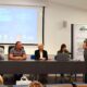 Panel od četvero ljudi koji sjede za stolom ispred ekrana s prezentacijom. Tema konferencije je "Sport i zdravlje", a jedan od govornika, žena, drži govor s govornice, dok ostali panelisti sjede i slušaju.