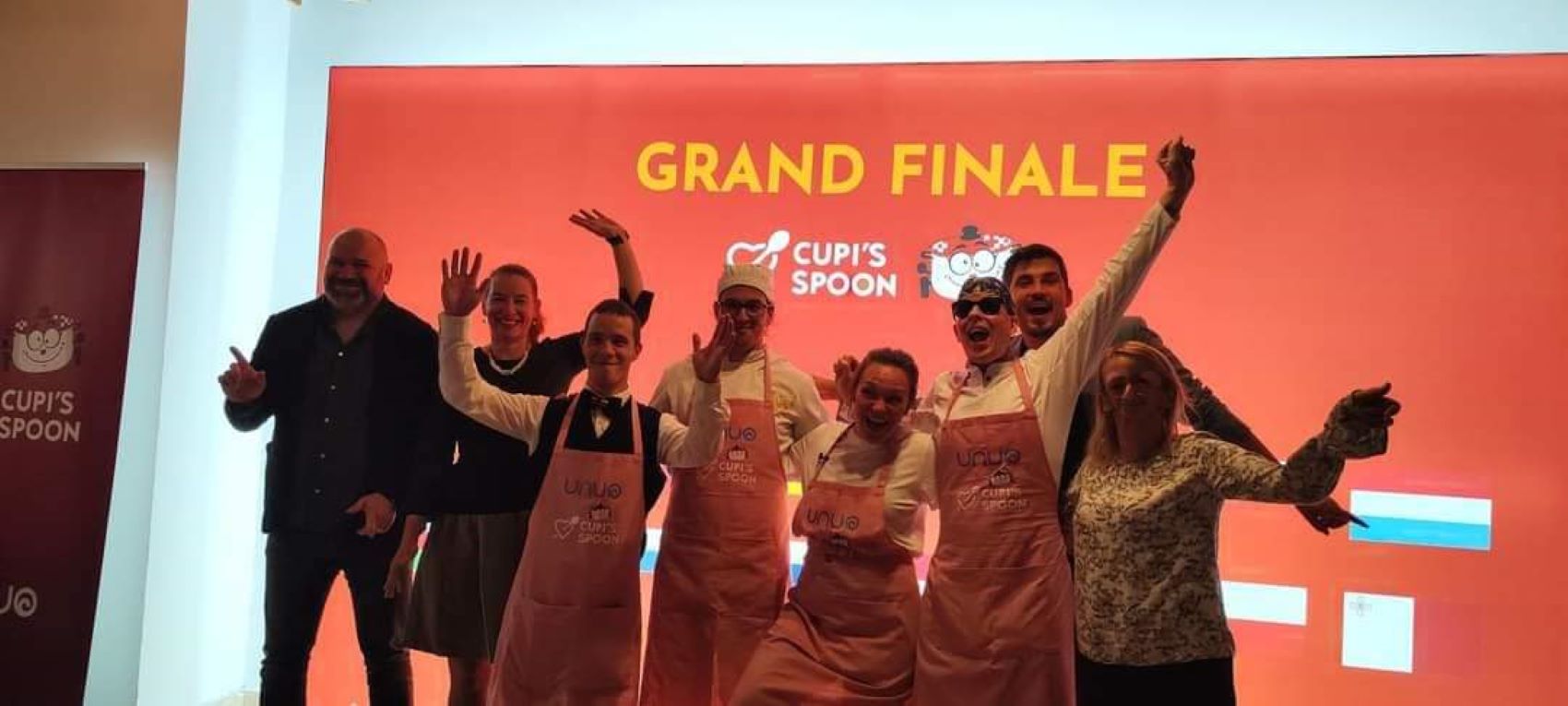 Na fotografiji je skupina ljudi koji poziraju ispred velikog natpisa “GRAND FINALE” na crvenoj pozadini s logotipom “CUP’S SPOON”. Neki od sudionika nose kuharske pregače s logotipom, što sugerira da se radi o kulinarskom događaju ili natjecanju.