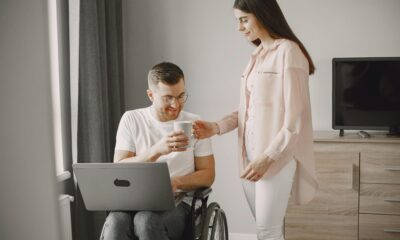 Fotografija prikazuje dvoje ljudi, muškaraca u invalidskim kolicima i ženu koja stoji pokraj njega i pruža mu bijelu šalicu. Muškarcu na koljenima stoji otvoren laptop. Iza njih nalazi se siva komoda na kojoj stoji televizija. Njih dvoje su dobro raspoloženi i uživaju u ugodnosti svoga doma.