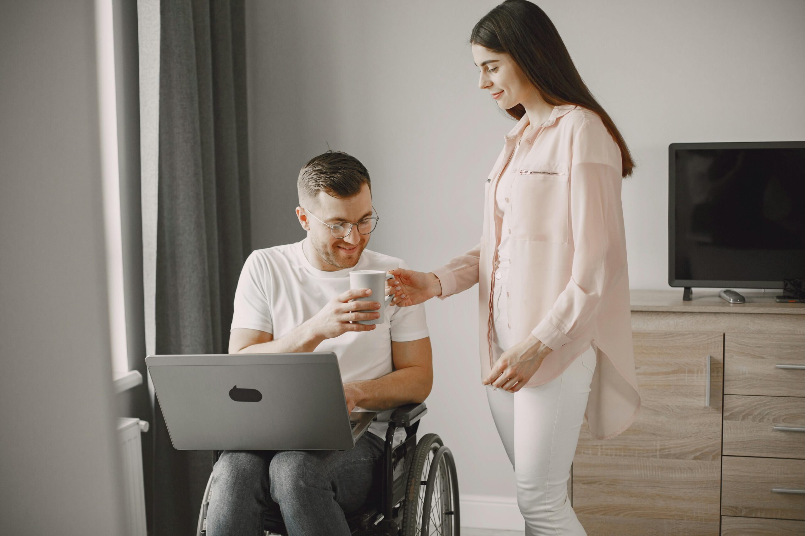 Fotografija prikazuje dvoje ljudi, muškaraca u invalidskim kolicima i ženu koja stoji pokraj njega i pruža mu bijelu šalicu. Muškarcu na koljenima stoji otvoren laptop. Iza njih nalazi se siva komoda na kojoj stoji televizija. Njih dvoje su dobro raspoloženi i uživaju u ugodnosti svoga doma.