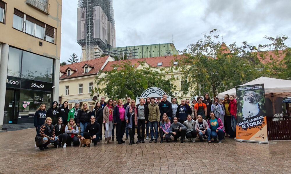 Skupina ljudi pozira za zajedničku fotografiju na gradskom trgu. Iza njih su zgrade i visoka građevina u obnovi, prekrivena skelama. U grupi su ljudi različite dobi, a svi izgledaju veselo i okupljeni su oko aktivnosti u vezi s udomljavanjem životinja, sudeći prema plakatu na desnoj strani s natpisom "Udomljavanje je fora!".