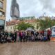 Skupina ljudi pozira za zajedničku fotografiju na gradskom trgu. Iza njih su zgrade i visoka građevina u obnovi, prekrivena skelama. U grupi su ljudi različite dobi, a svi izgledaju veselo i okupljeni su oko aktivnosti u vezi s udomljavanjem životinja, sudeći prema plakatu na desnoj strani s natpisom "Udomljavanje je fora!".