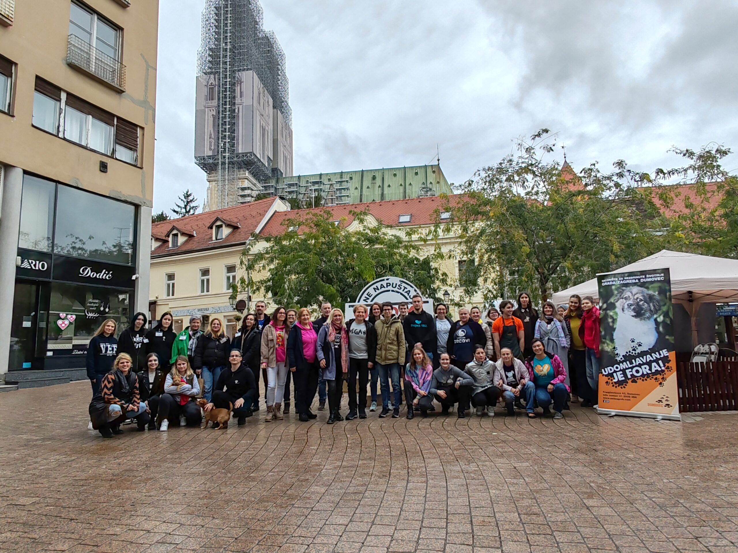 Skupina ljudi pozira za zajedničku fotografiju na gradskom trgu. Iza njih su zgrade i visoka građevina u obnovi, prekrivena skelama. U grupi su ljudi različite dobi, a svi izgledaju veselo i okupljeni su oko aktivnosti u vezi s udomljavanjem životinja, sudeći prema plakatu na desnoj strani s natpisom "Udomljavanje je fora!".