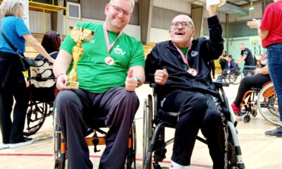 Fotografija prikazuje dvije osobe u invalidskim kolicima, jedna osoba drži pehar i oko vrata nosi medalju, dok druga ima samo medalju oko vrata. Jedan muškarac je obučen u zelenu majicu i crnu trenirku, dok drugi ima crnu majicu i crnu trenirku. U daljini slike vide se boće što znači da je riječ o boćarskom turniru.