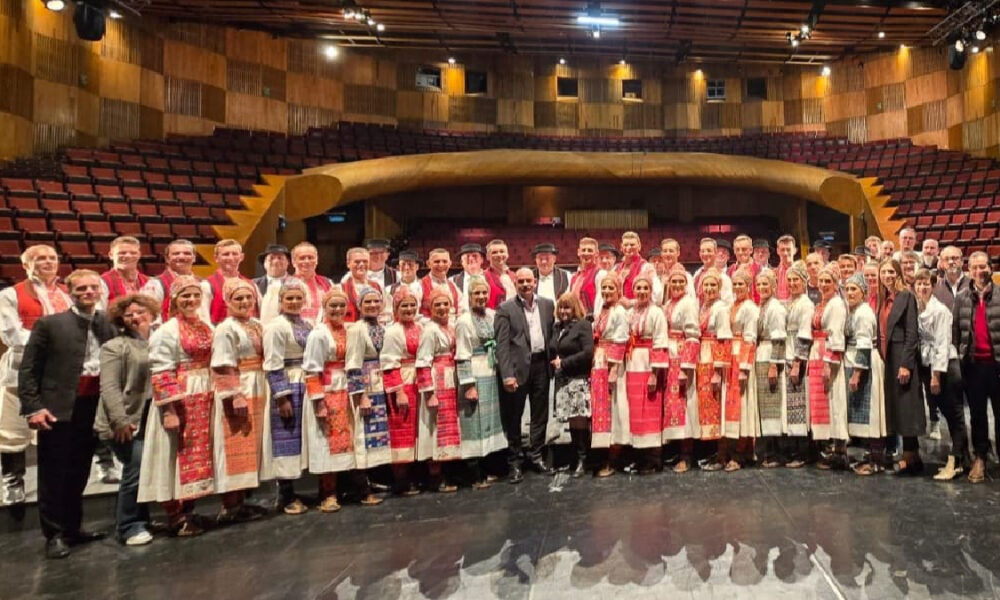 Na slici je velika skupina ljudi u narodnim nošnjama, članovi ansambla LADO, koji poziraju na pozornici nakon izvedbe. Svi su odjeveni u tradicionalne hrvatske narodne nošnje i stoje ispred praznog gledališta u velikoj koncertnoj dvorani, što sugerira završetak kulturnog događaja ili koncerta.