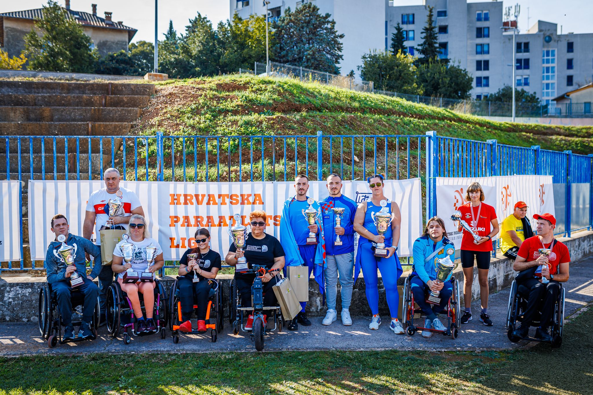 Na slici je grupa sportaša s invaliditetom okupljenih ispred natpisa "Hrvatska paraatletska liga". Neki su u kolicima, a u rukama drže velike pehare, ponosno pokazujući svoje nagrade. Pozadina prikazuje vanjski sportski teren okružen plavom ogradom, a iza toga se vide stambene zgrade. Atmosfera na slici odiše ponosom i zajedništvom, a sportaši su obučeni u različite sportske odore, simbolizirajući uspjeh i postignuća u paraatletici.