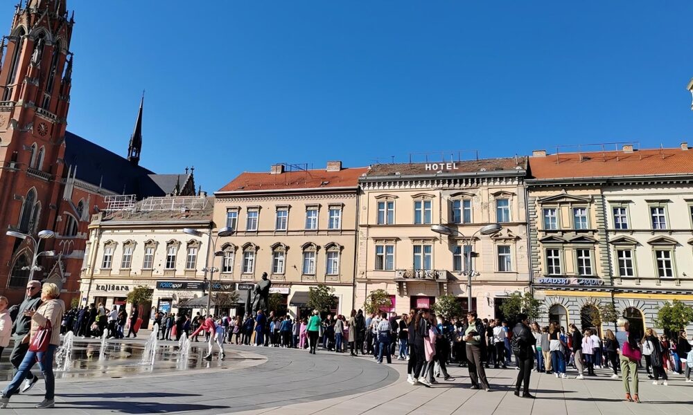 Slika prikazuje ljude koji se okupljaju na gradskom trgu ispred zgrade s povijesnom arhitekturom. Iza se vidi crkveni toranj, a na trgu su fontane. Okupljena grupa ljudi čeka ili sudjeluje u nekom događaju, a dan je sunčan, s vedrim plavim nebom.