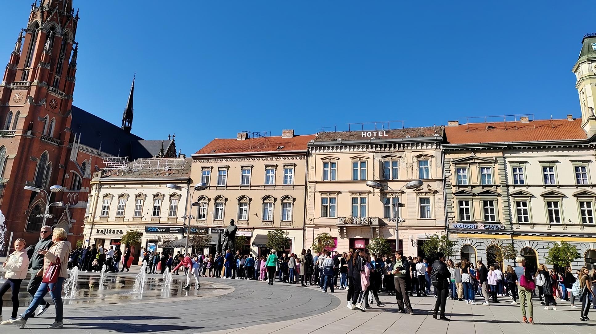 Slika prikazuje ljude koji se okupljaju na gradskom trgu ispred zgrade s povijesnom arhitekturom. Iza se vidi crkveni toranj, a na trgu su fontane. Okupljena grupa ljudi čeka ili sudjeluje u nekom događaju, a dan je sunčan, s vedrim plavim nebom.