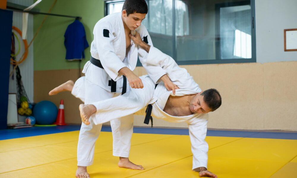 Fotografija prikazuje dva taekwondoista kako se pore na tatamiju. Lijevi tekwondoist ruši desnoga, dok se ovaj prima rukom za tatami. Podloga na kojoj se bore je žuto-plave boje.