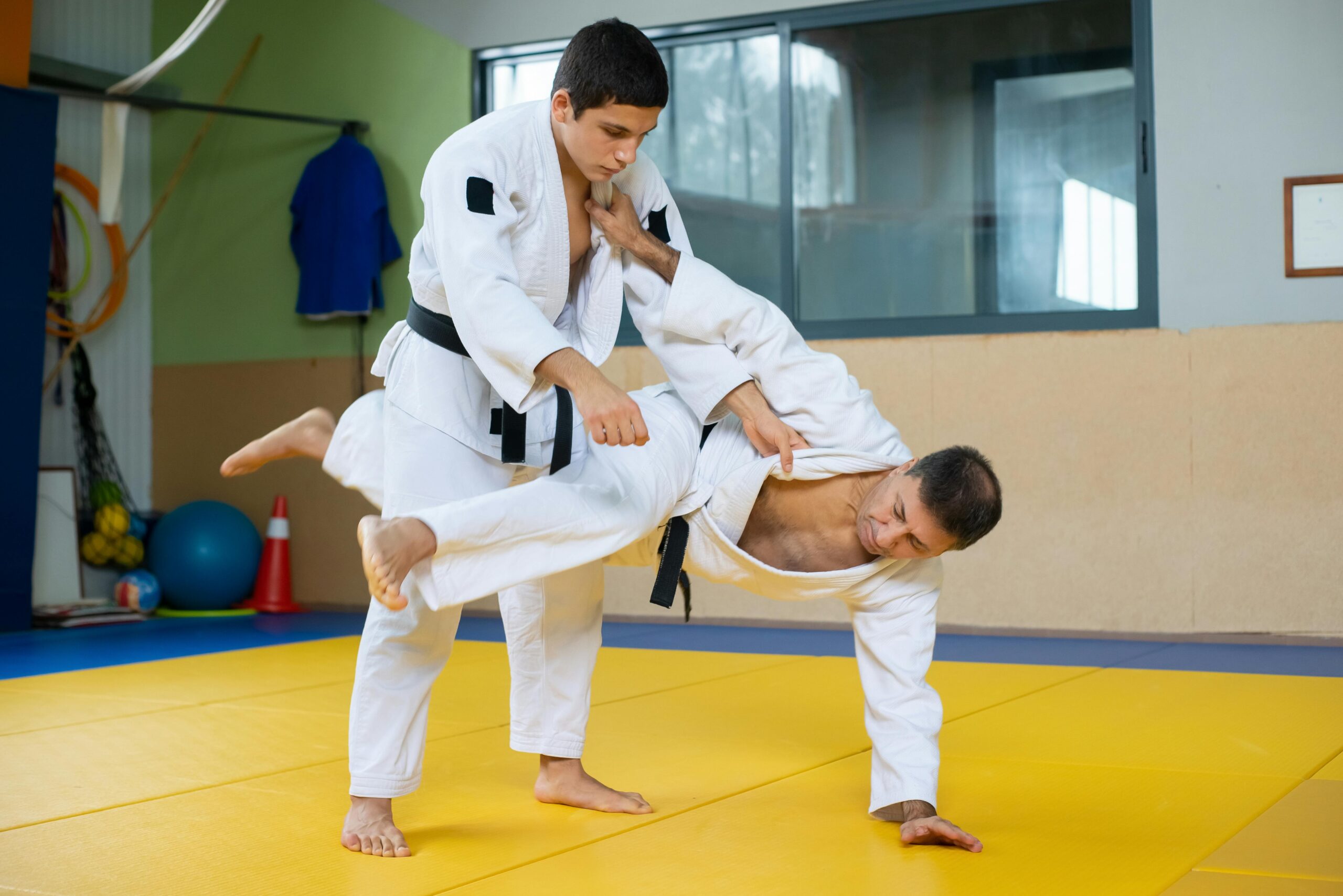 Fotografija prikazuje dva taekwondoista kako se pore na tatamiju. Lijevi tekwondoist ruši desnoga, dok se ovaj prima rukom za tatami. Podloga na kojoj se bore je žuto-plave boje.