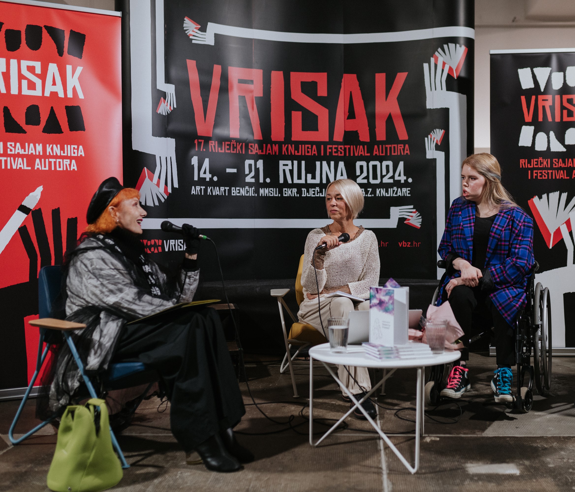 Fotografija prikazuje predstavljanje knjige poezije Branke Kostić. Četiri osobe sjede ispred pozadine s velikim plakatima na kojima piše “VRISAK” i informacije o 17. Riječkom sajmu knjiga i festivalu autora koji se održava od 14. do 21. rujna 2024. godine.