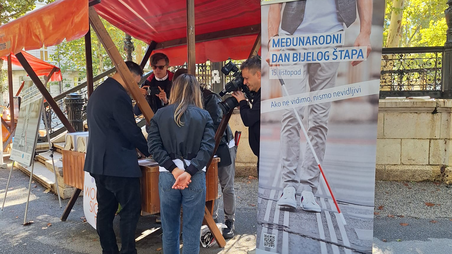 Ova slika prikazuje promotivni štand postavljen na otvorenom povodom Međunarodnog dana bijelog štapa, 15. listopada. Nekoliko osoba sudjeluje u događaju ispod crvenih nadstrešnica, dok fotografi snimaju prizor. Na plakatu s desne strane stoji natpis "Međunarodni dan bijelog štapa" uz poruku "Ako ne vidimo, nismo nevidljivi."