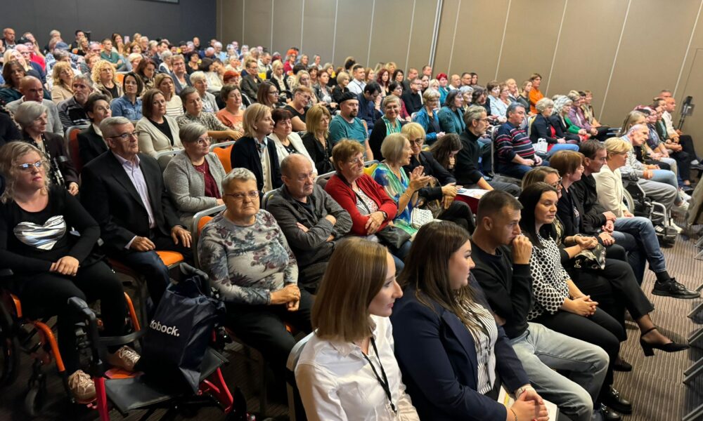 Slika prikazuje veliku publiku u zatvorenom prostoru. Većinom stariji ljudi sjede u redovima, pažljivo slušajući ili promatrajući događaj ispred njih. U prednjem planu nalaze se osobe koje koriste hodalicu, a atmosfera je ozbiljna i fokusirana.