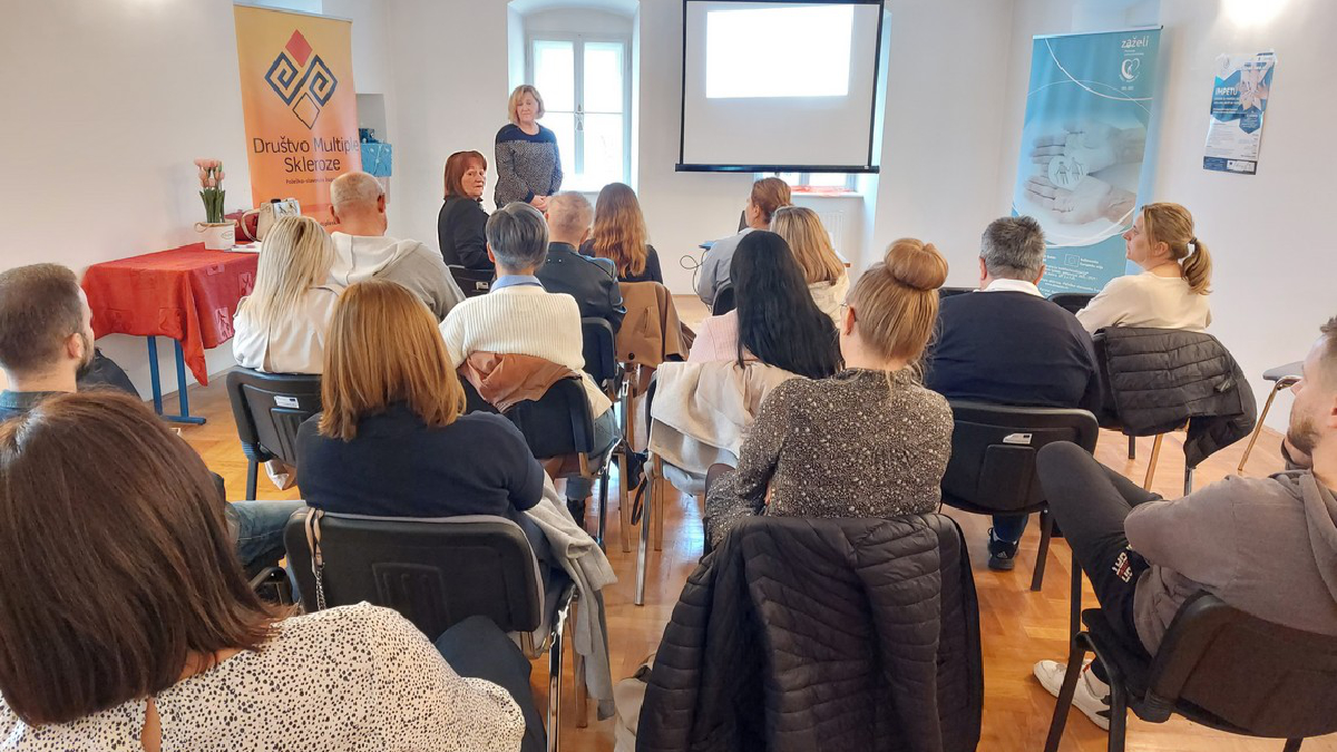 Skupina ljudi sjedi u prostoriji u kojoj se održava predavanje ili prezentacija. Osoba stoji ispred publike, pored projektora koji prikazuje prezentaciju na platnu. Sudionici su okrenuti prema predavačici, pažljivo slušajući. U pozadini s lijeve strane nalazi se plakat s logotipom i natpisom "Društvo multiple skleroze", dok je desno postavljen promotivni banner s informacijama o nekom programu ili projektu. Atmosfera je neformalna, a prostorija svijetla, s velikim prozorom u pozadini.