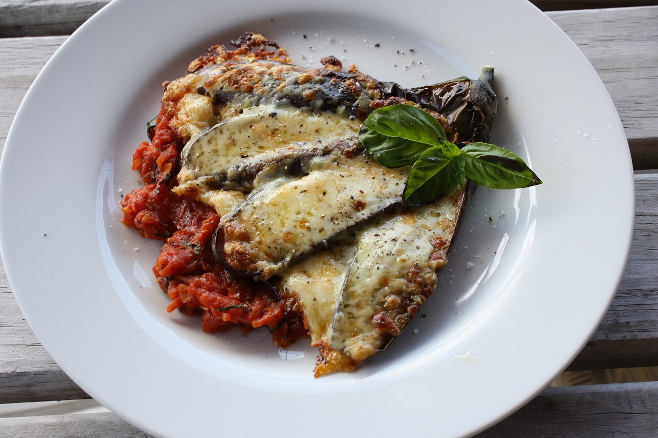 Na fotografiji je prikazan tanjur s hranom koji sadrži jelo koje izgleda kao parmigiana di melanzane, talijansko jelo od patlidžana. Jelo se sastoji od slojeva pečenog patlidžana i umaka od rajčice, prekriveno rastopljenim sirom i začinjeno. Na vrhu jela nalazi se list bosiljka koji dodaje svježinu i boju. Fotografija bi mogla biti zanimljiva ili relevantna zbog prikaza tradicionalnog talijanskog jela koje je popularno u mnogim kulturama, a može biti korisno za ljude koji traže inspiraciju za kuhanje ili žele znati više o različitim kuhinjama svijeta.