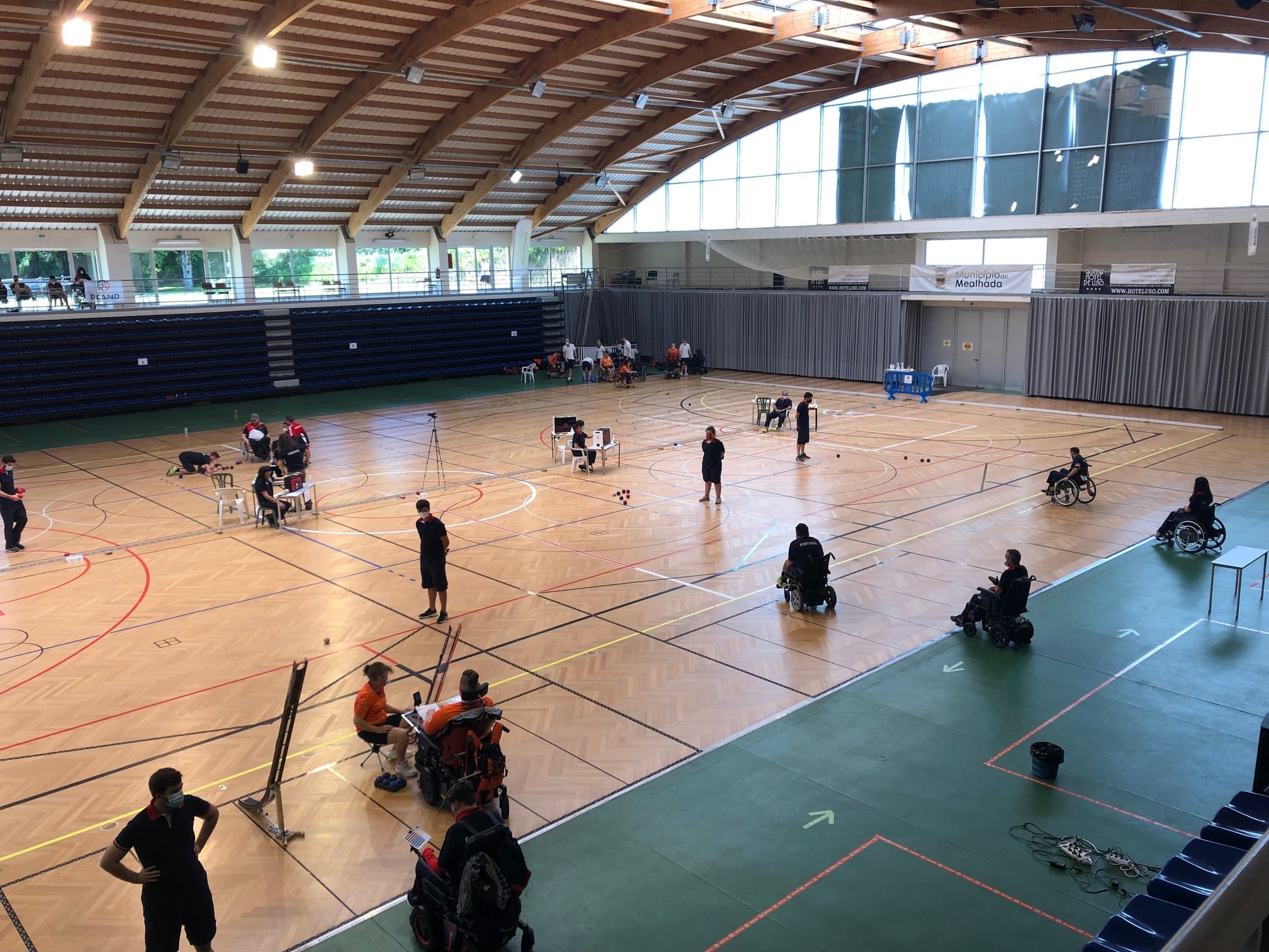 Fotografija prikazuje natjecanje u bocci. Više ljudi u kolicima igra na više polja. Natjecanje se održalo u sportskoj dvorani.