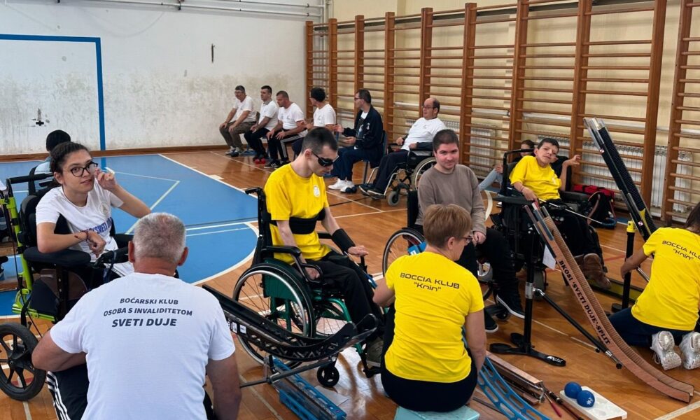 Fotografija prikazuje nekoliko boćara u kolicima i nekoliko njih koji sjede na klupi. Riječ je o bocci. Natjecanje se održalo u sportskoj dvorani što se da primijetiti po košu, golu i švedskim ljestvama. Neki od boćara boćaju uz pomoć asistenta i rampe.