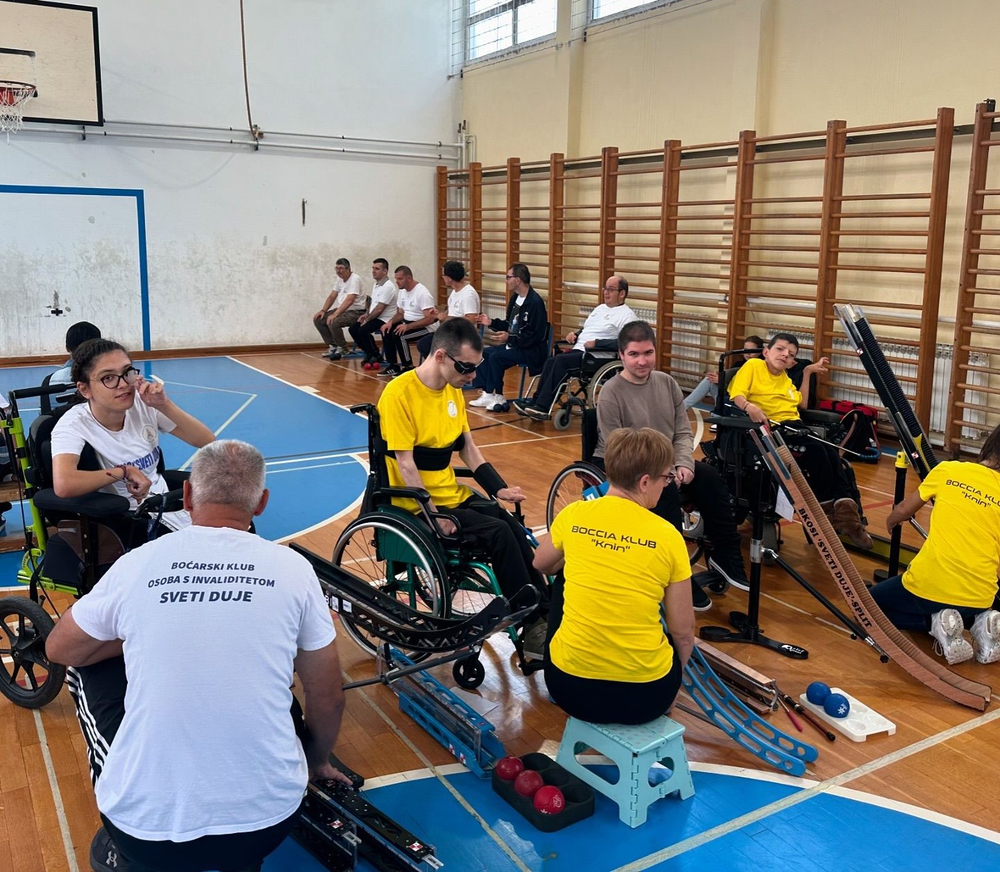 Fotografija prikazuje nekoliko boćara u kolicima i nekoliko njih koji sjede na klupi. Riječ je o bocci. Natjecanje se održalo u sportskoj dvorani što se da primijetiti po košu, golu i švedskim ljestvama. Neki od boćara boćaju uz pomoć asistenta i rampe.