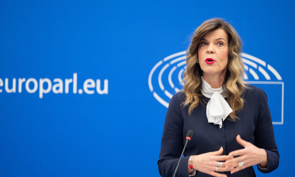 Žena govori ispred plave pozadine s natpisom "europarl.eu" i logotipom Europskog parlamenta. Ima dugu, valovitu kosu i nosi tamnu bluzu s bijelom mašnom. Gestikulira rukama i čini se da se obraća publici. Ispred nje na postolju nalazi se mikrofon.