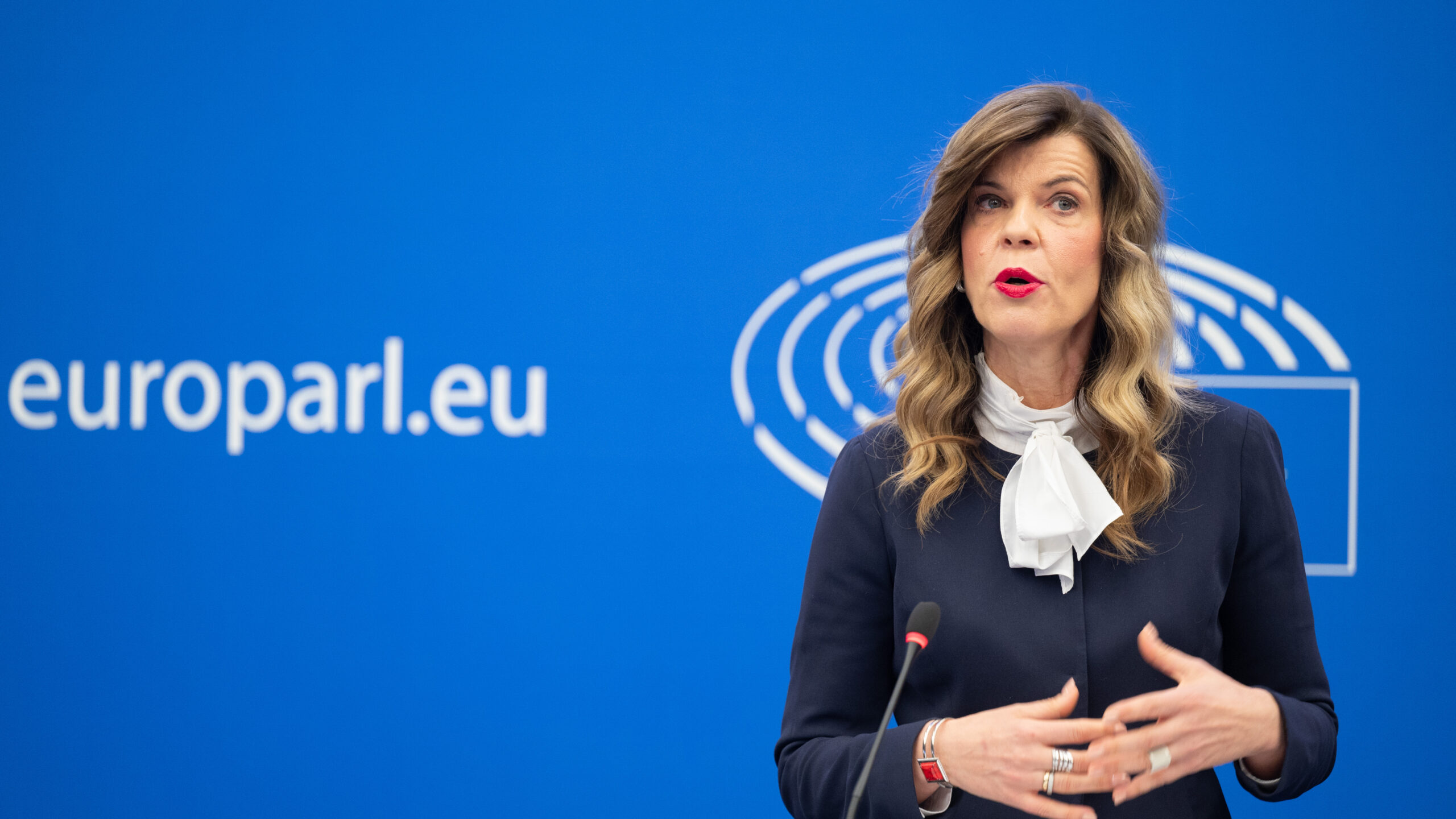 Žena govori ispred plave pozadine s natpisom "europarl.eu" i logotipom Europskog parlamenta. Ima dugu, valovitu kosu i nosi tamnu bluzu s bijelom mašnom. Gestikulira rukama i čini se da se obraća publici. Ispred nje na postolju nalazi se mikrofon.