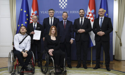 Grupna fotografija snimljena ispred plavog službenog bannera s grbom Republike Hrvatske i natpisom "VLADA REPUBLIKE HRVATSKE". Na slici je osmero ljudi – dvoje u invalidskim kolicima u prvom planu i šestorica stojeći iza njih. Dva zastavna jarbola sa zastavama Hrvatske i Europske unije nalaze se s obje strane fotografije. Atmosfera je formalna, a sudionici izgledaju zadovoljno i profesionalno.