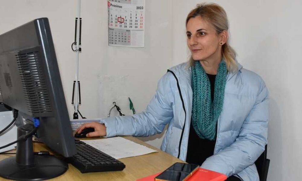 Na fotografiji je osoba koja sjedi za stolom ispred računala. Osoba nosi svijetloplavu jaknu i zeleni šal. Na stolu se nalazi crni monitor, tipkovnica, miš, bijeli uređaj (vjerojatno modem ili router), te crveni fascikl i mobitel. Na zidu iza osobe nalazi se kalendar za prosinac 2023. godine.