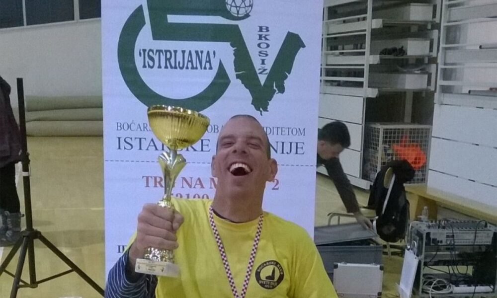 Na fotografiji se nalazi osoba u invalidskim kolicima koja drži zlatni pehar u desnoj ruci. Osoba nosi žutu majicu s logom na lijevoj strani prsa i medalju oko vrata. Iza osobe je plakat s natpisom "ISTRIJANA" i logom koji prikazuje invalidska kolica s loptom. Na plakatu također piše "BOĆARSKI KLUB OSOBA S INVALIDITETOM ISTARSKE ŽUPANIJE". U pozadini se vidi unutrašnjost sportske dvorane s raznim tehničkim uređajima i opremom.